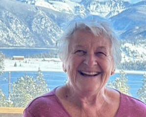 An elderly person smiling with a snowy mountain and lake landscape in the background.