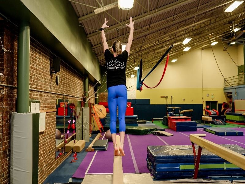 A gymnast balances on a beam with arms raised in a gym.