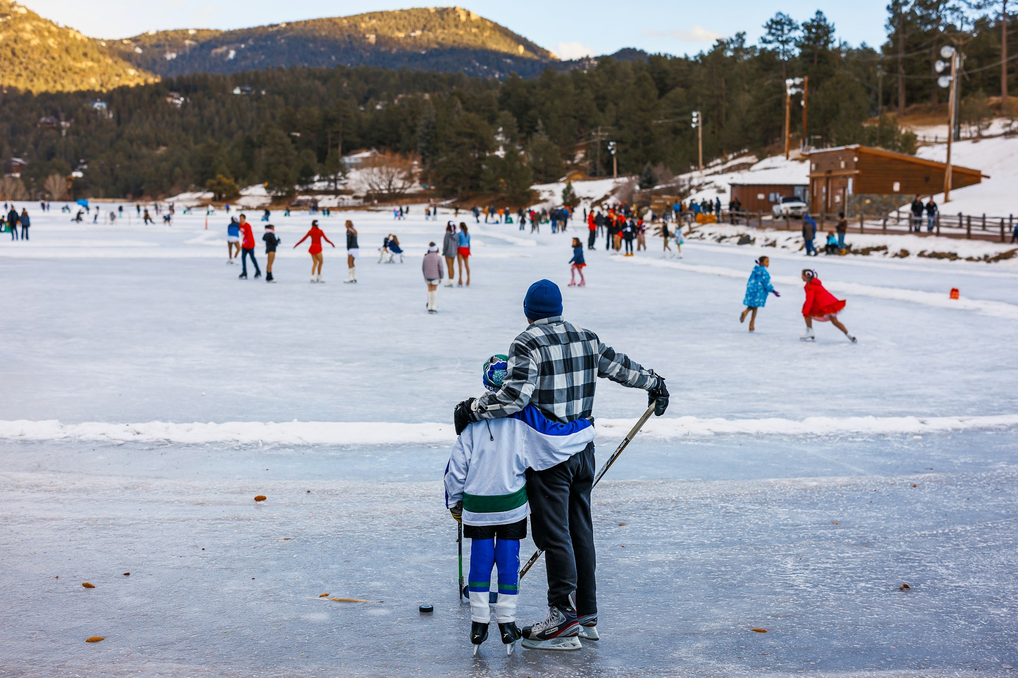 People skating on an outdoor ice rink, surrounded by snowy landscape and trees; a pair stands at the front holding hockey sticks.