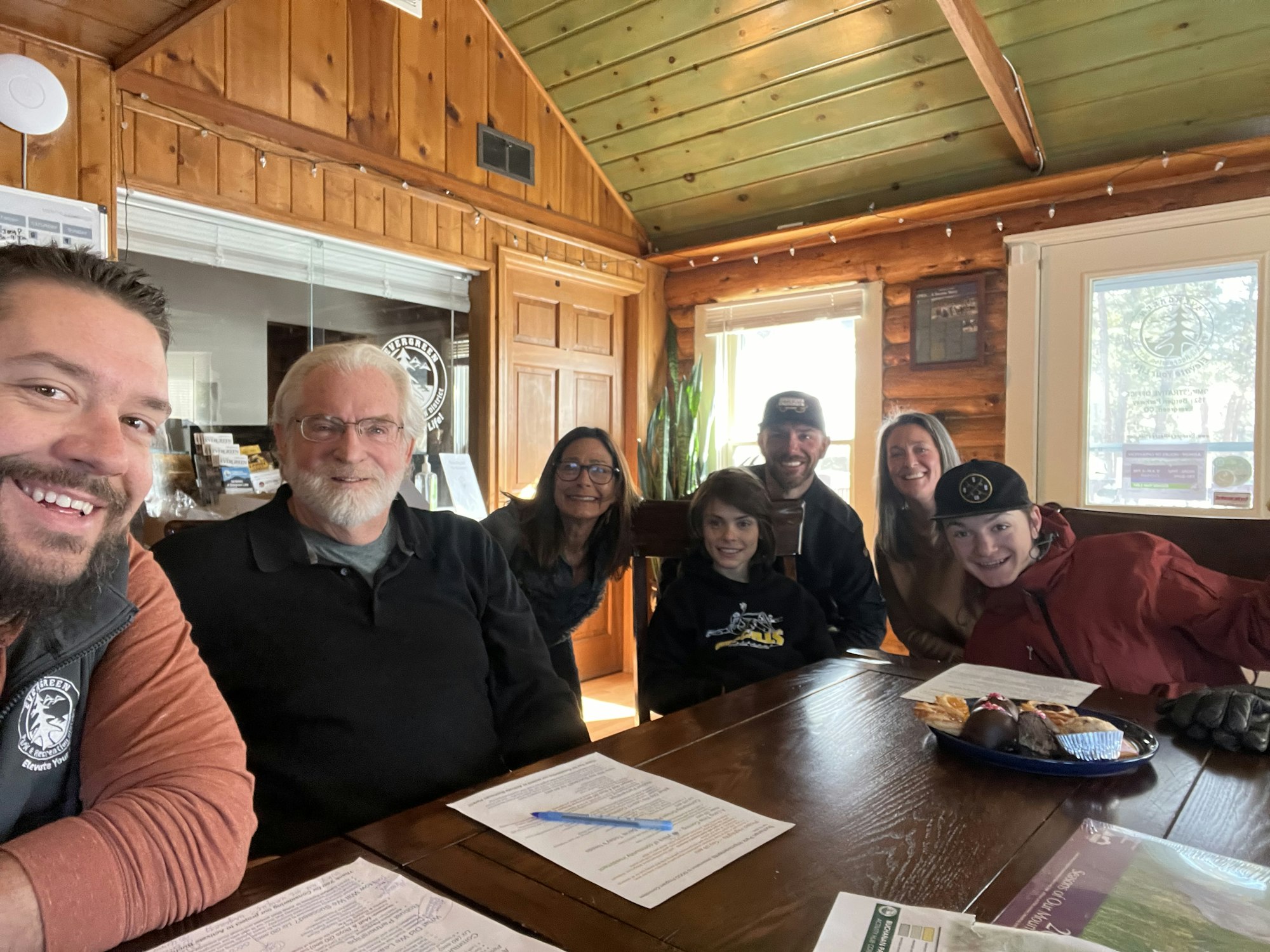 A group of people seated at a wooden table, with documents and snacks, in a cozy cabin-like setting.