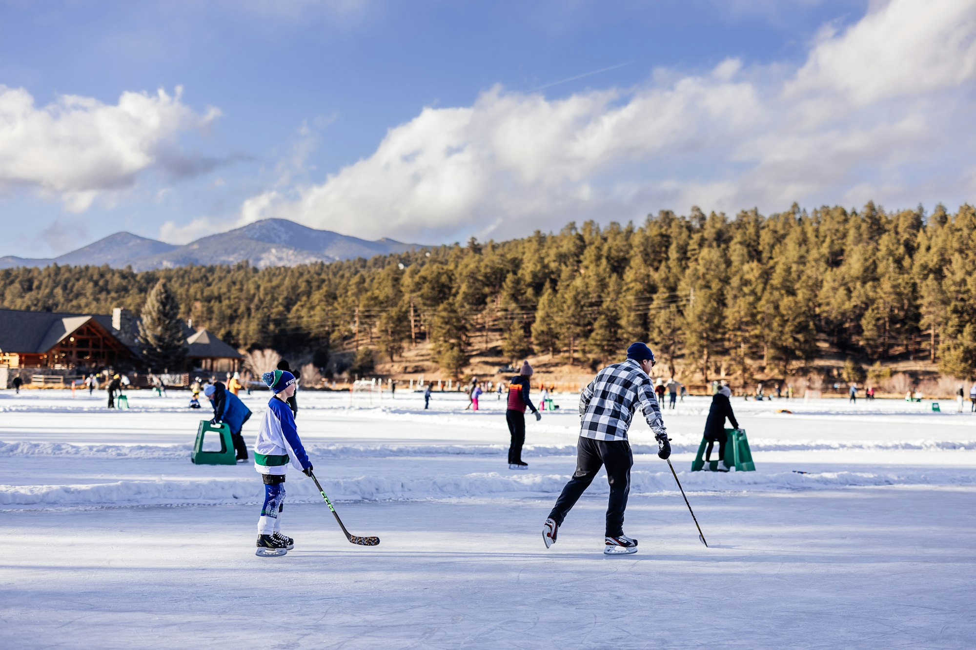 People playing ice hockey on a frozen lake, surrounded by trees and mountains, under a partly cloudy sky.