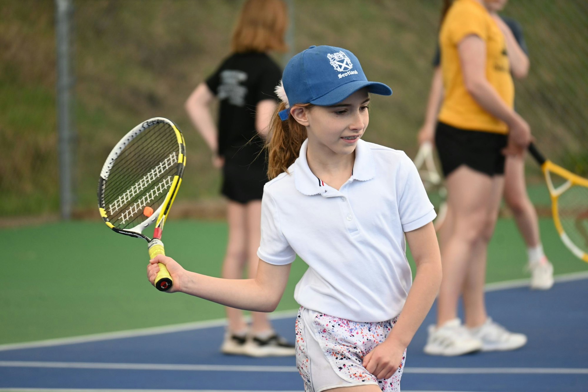 A young girl with a cap holding a tennis racket on a court, with blurred players in the background.