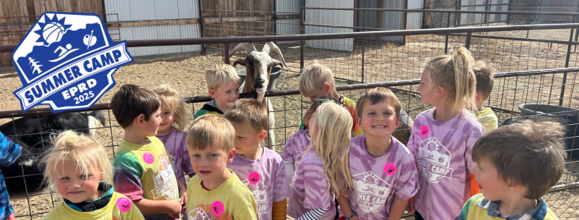 A group of children in colorful shirts at a summer camp with goats in a pen.