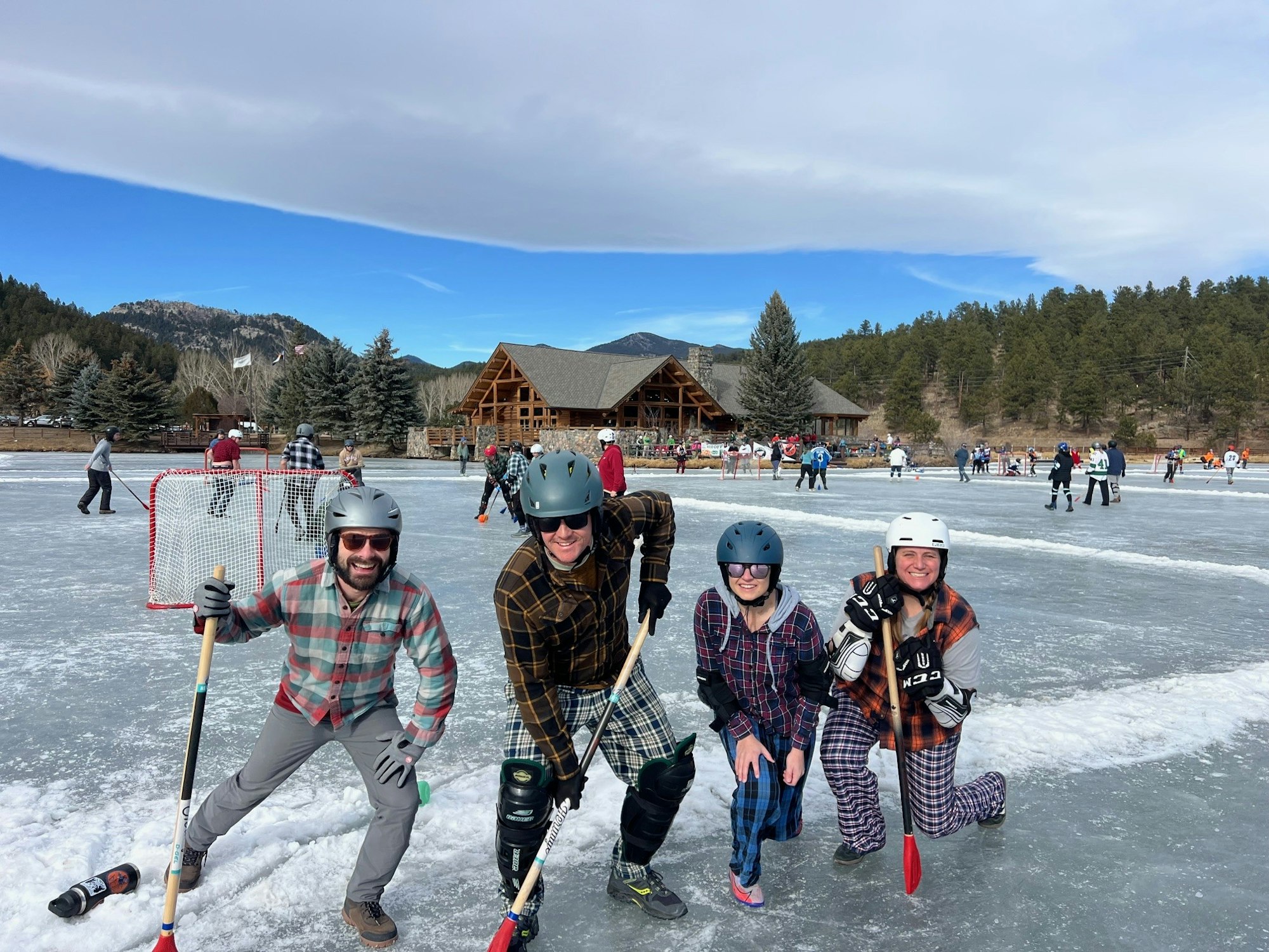 A group of four people in helmets and plaid outfits pose on an ice rink, while others play hockey in the background.