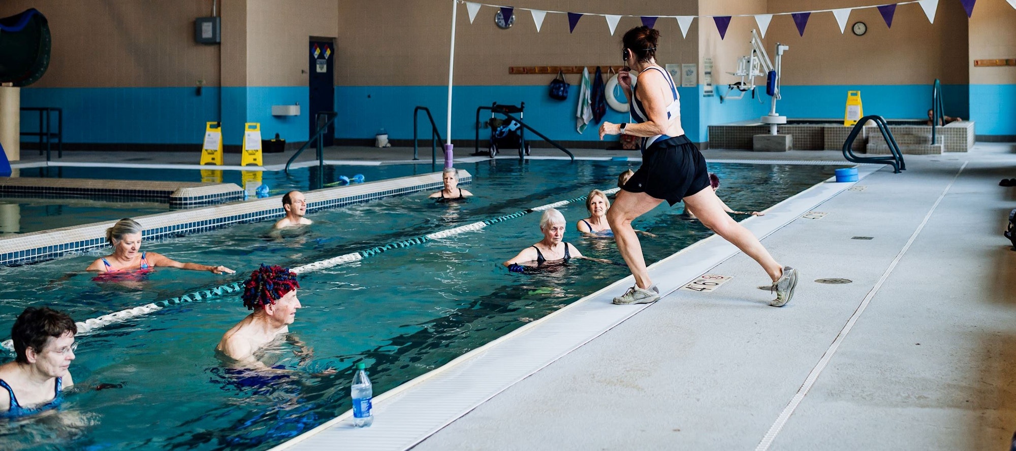 An indoor pool with people exercising in water and one person running by the poolside.