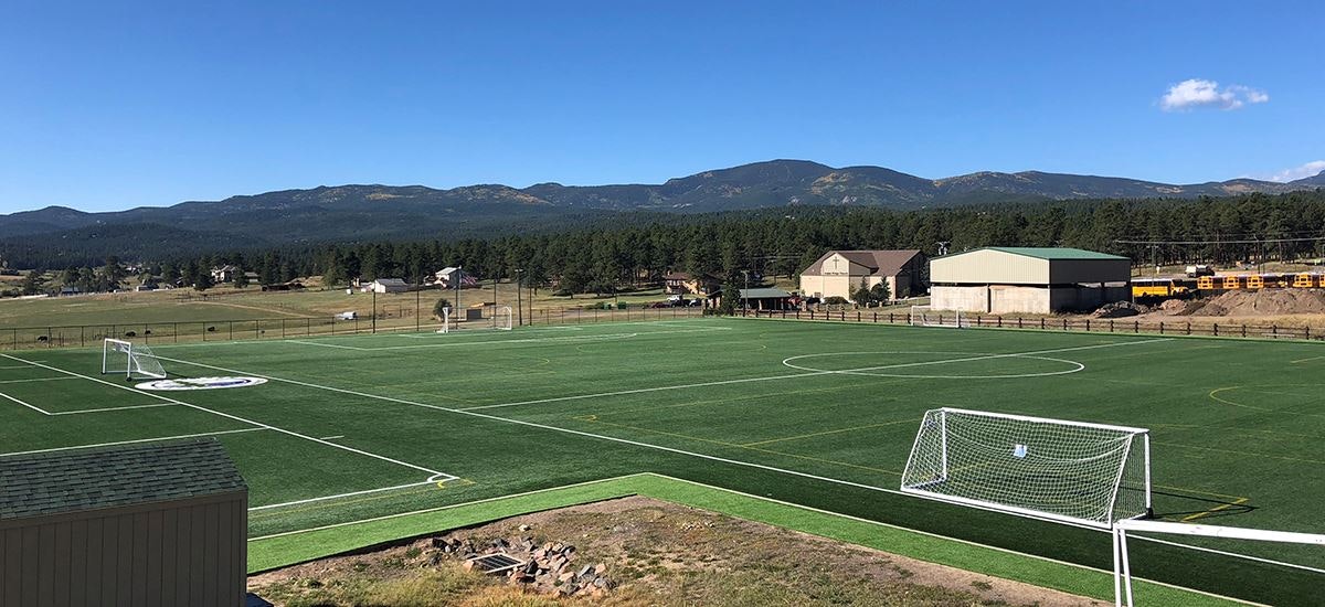 An empty soccer field with artificial turf, mountains in the background, and clear blue skies.