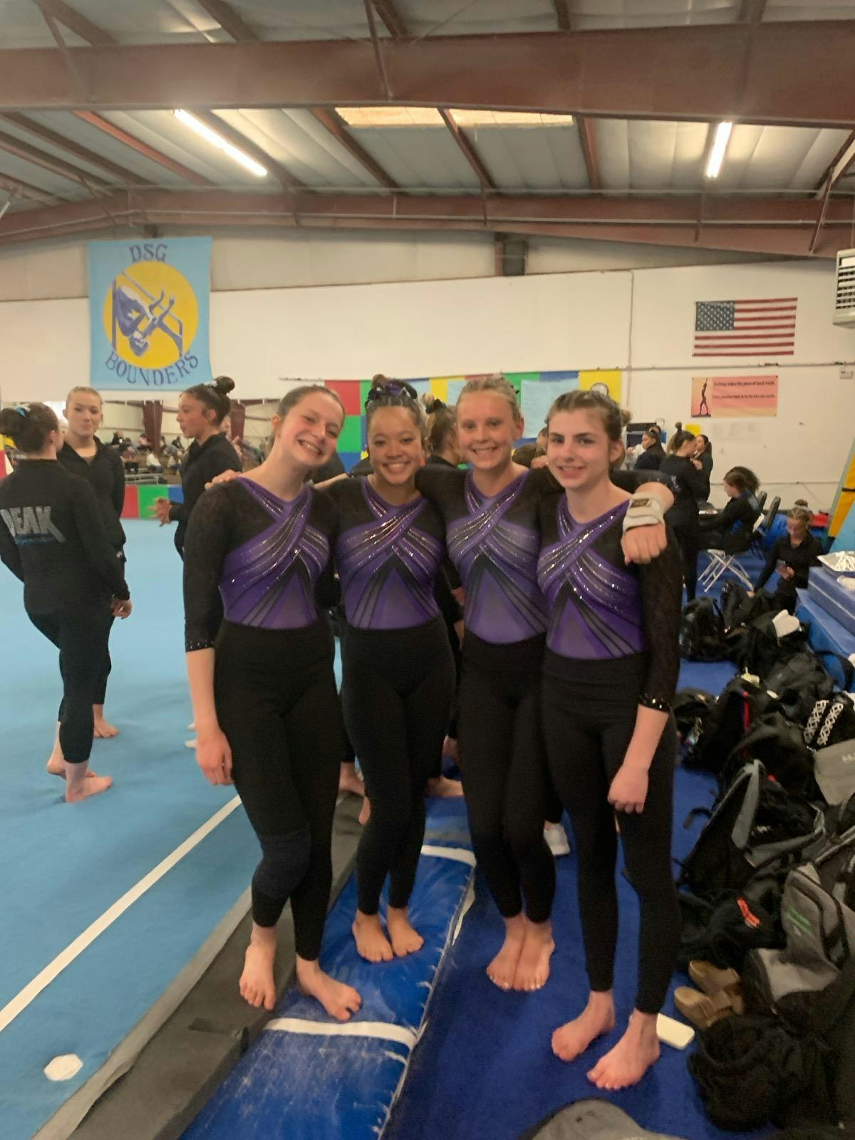 Four gymnasts in matching purple attire pose together in an indoor gym.