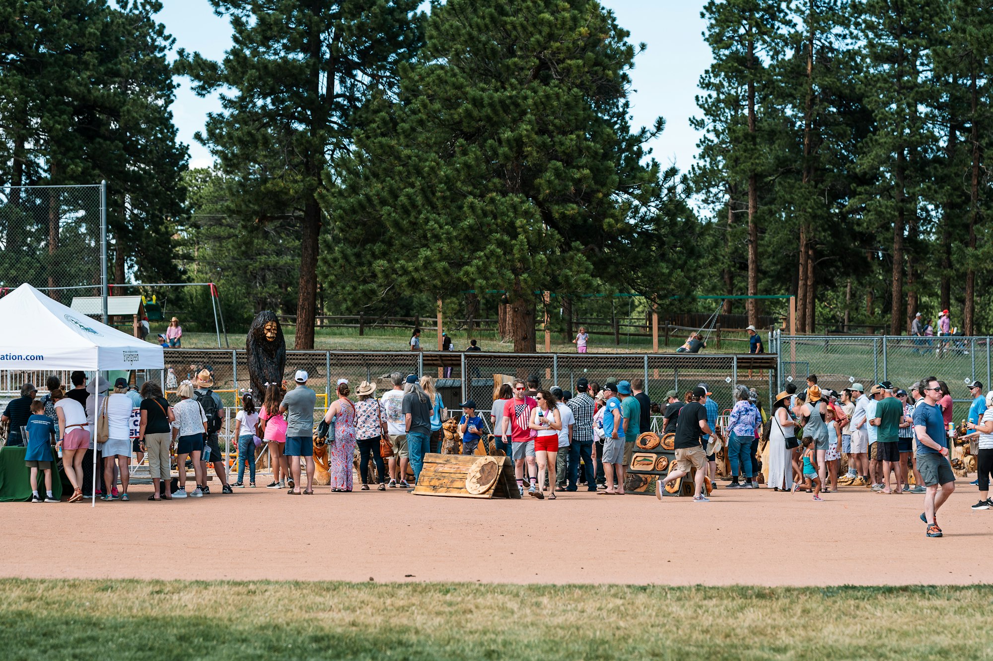 A crowd gathered at an outdoor event with tents, wooden carvings, and tall trees in the background.