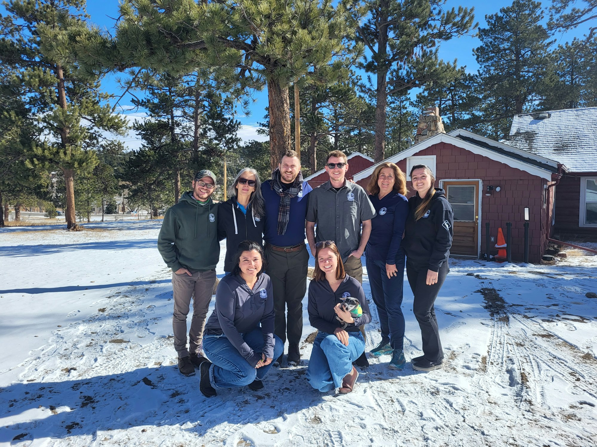 A group of people posing outdoors in the snow with trees and a cabin in the background.