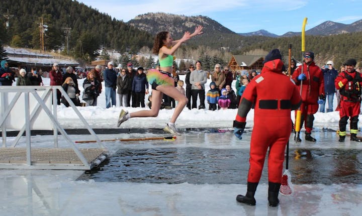 A person in colorful swimwear jumps into icy water, while onlookers and rescue personnel watch. It looks like a chilly event!
