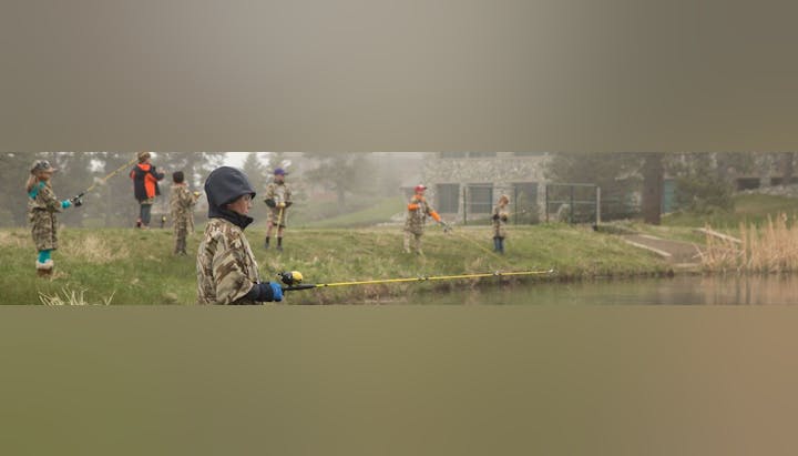 Children in camouflage outfits fishing by a pond, with foggy background and a building on the right.