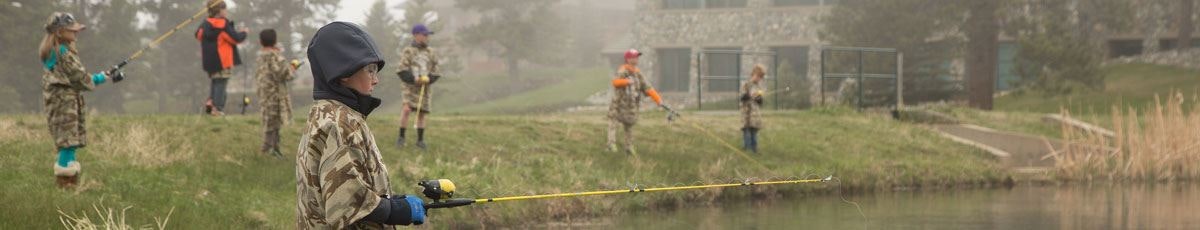 Children in camouflage outfits fishing by a pond, with foggy background and a building on the right.