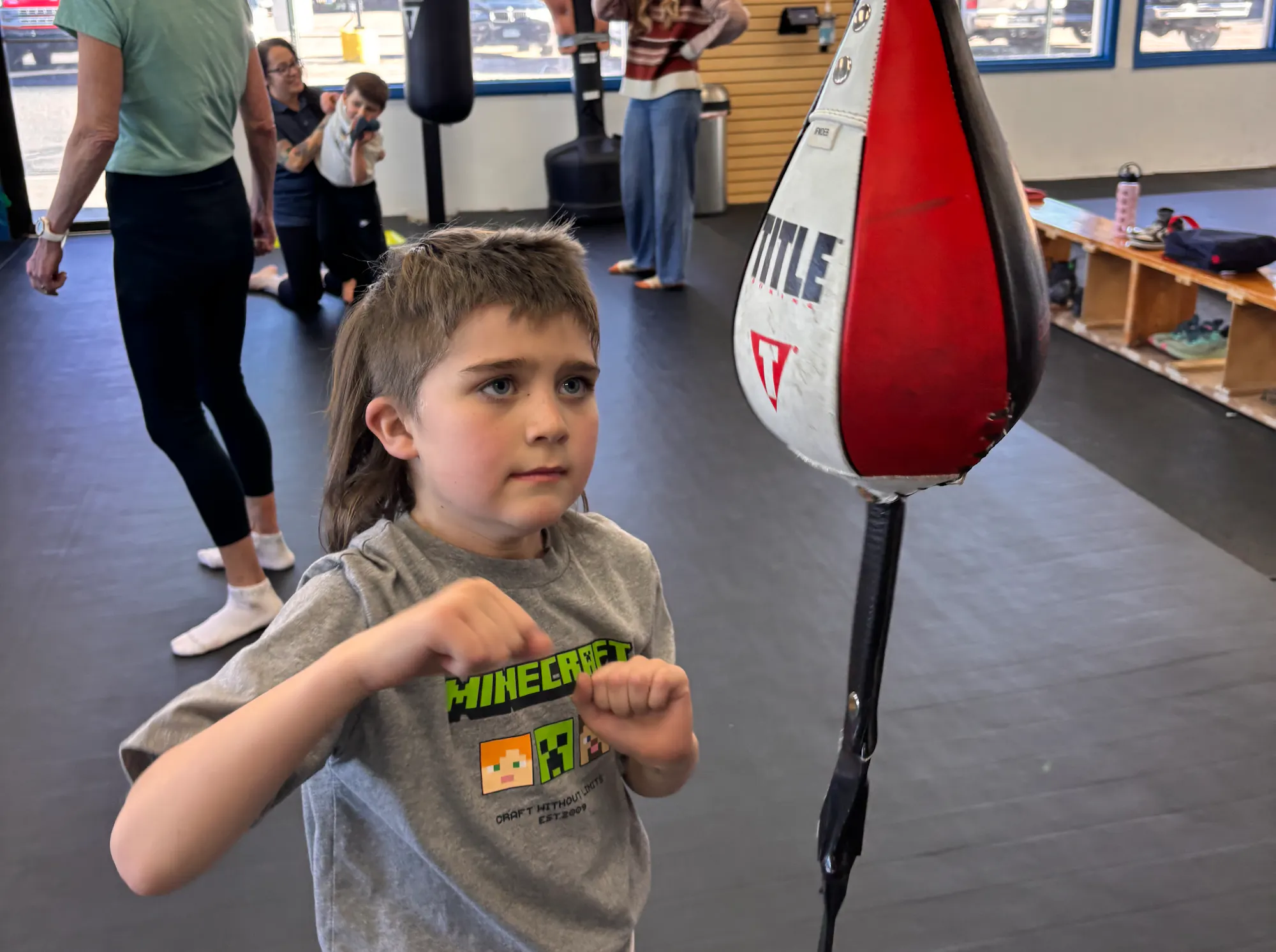 A child in a gym is practicing boxing with a punching bag, while others are engaged in different activities in the background.