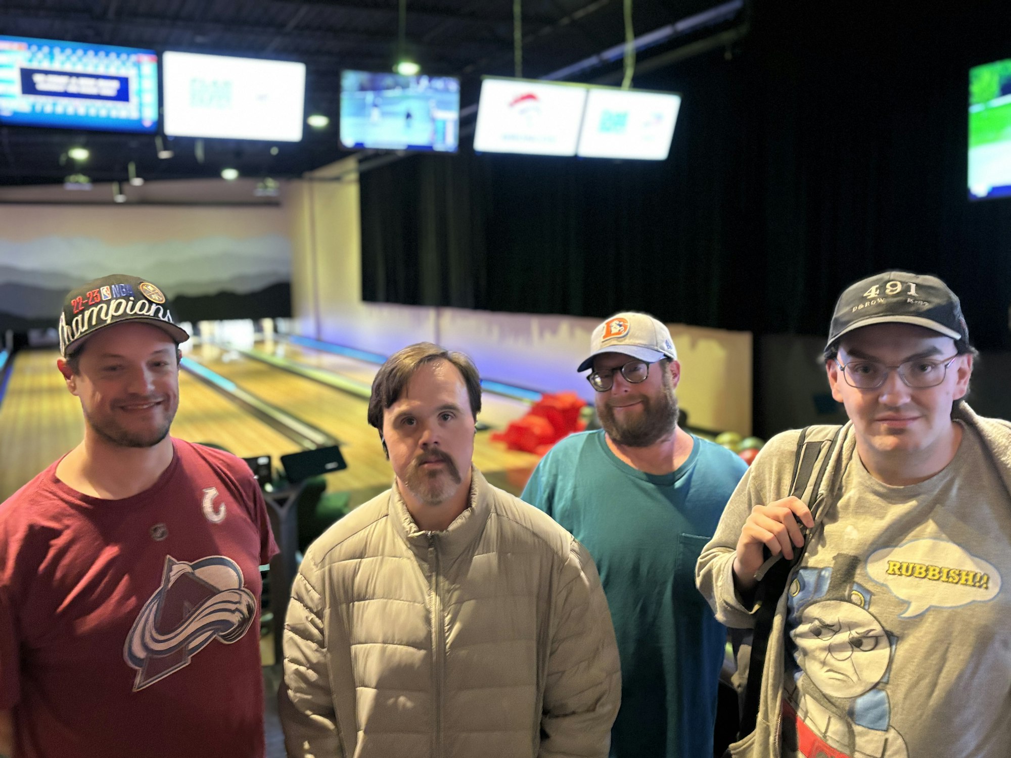 Four people standing in a bowling alley, smiling; bowling lanes and screens in the background.