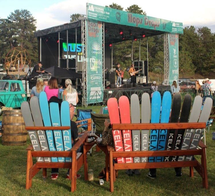 Outdoor concert with audience, colorful Adirondack chairs in foreground, band on stage, and signage indicating a sponsorship by "Hope Brew".