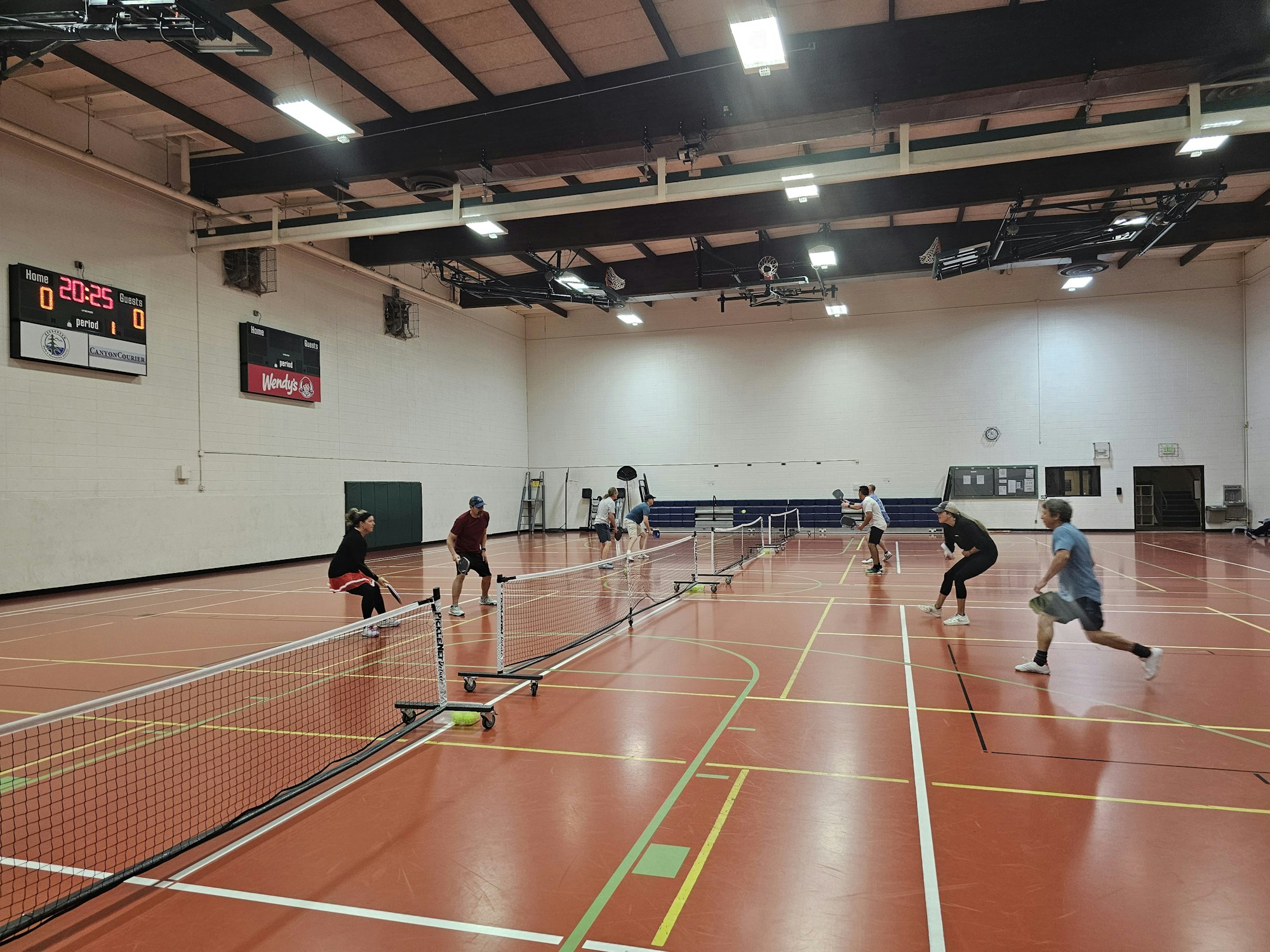 A group of people playing pickleball indoors in a gymnasium with red flooring and scoreboard visible.