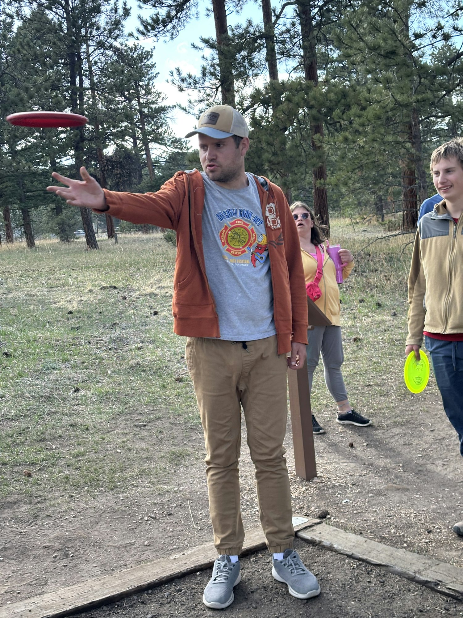People playing disc golf in a forested area, with a frisbee in motion.