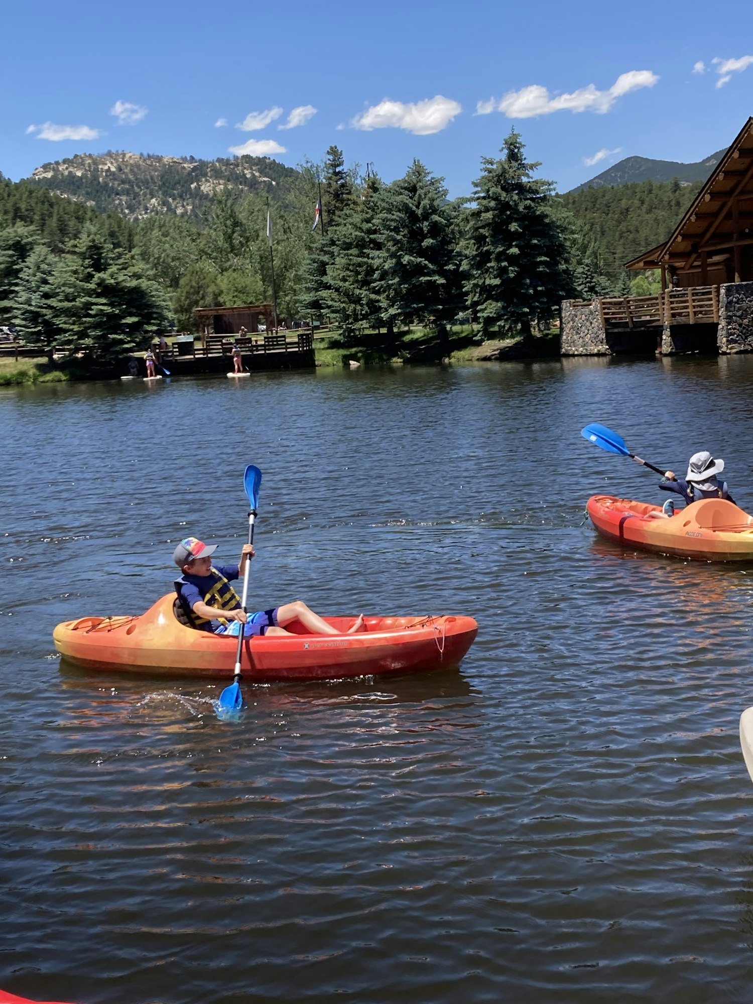 Two people kayaking on a lake near a forested shore and cabin, under a clear blue sky.