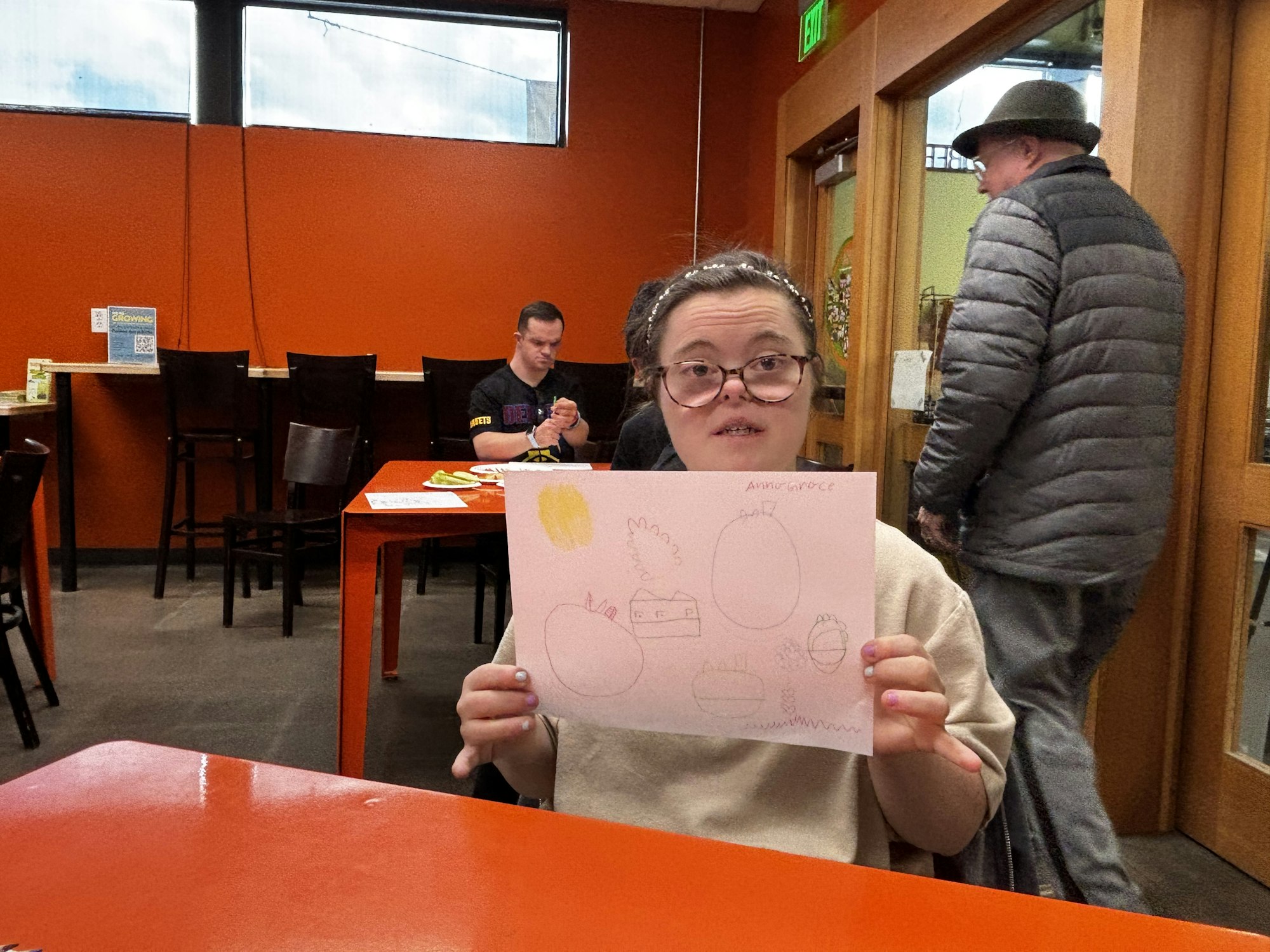A person holds a drawing in a room with orange tables and walls; another person sits at a table using their phone.