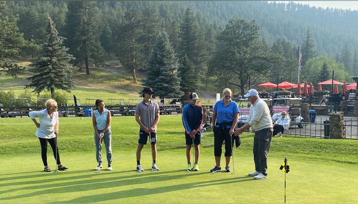 A group of people receiving golf instruction on a putting green, with carts and trees in the background.
