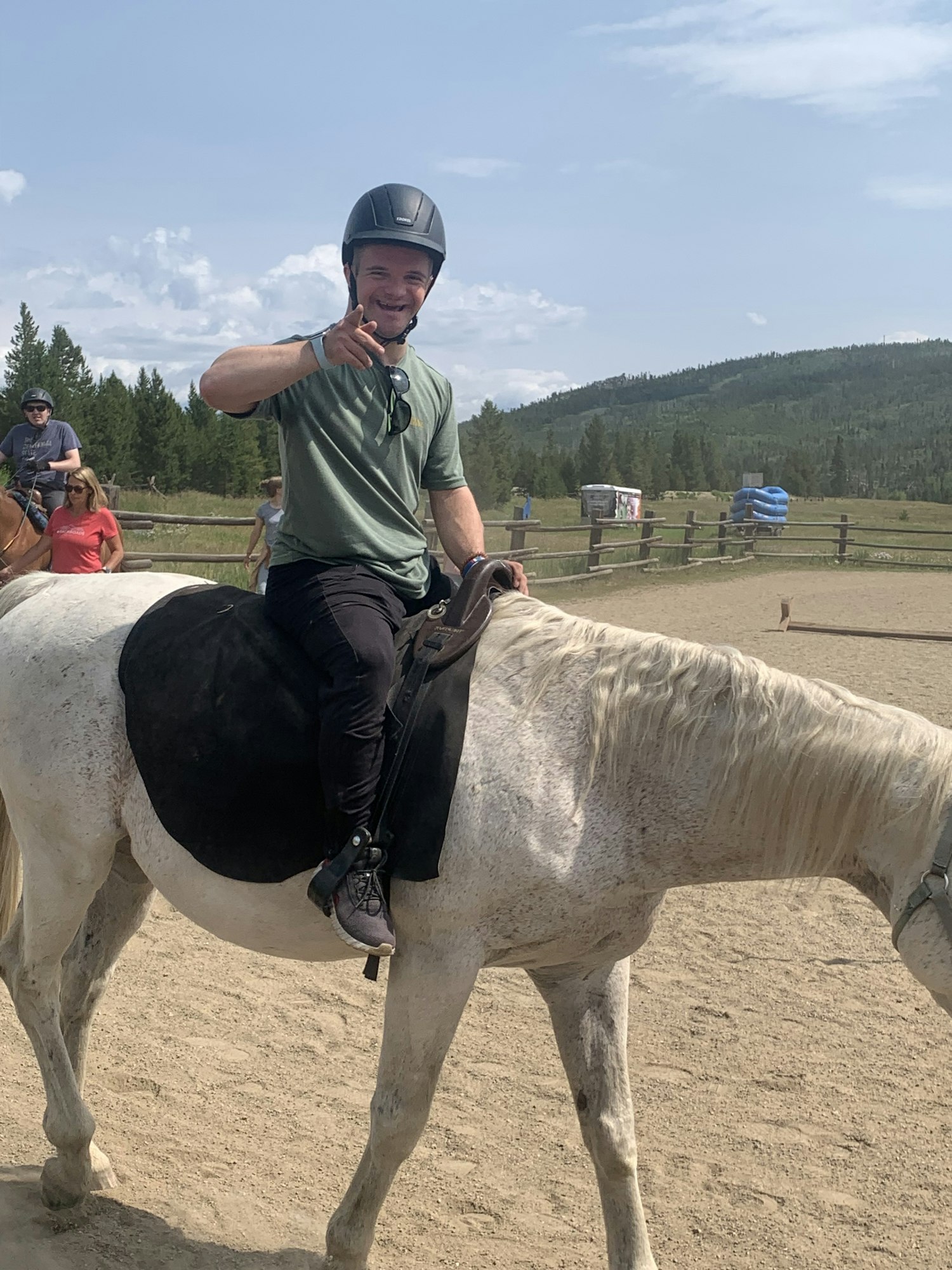 Person riding a white horse on a sandy area, wearing a helmet, with a forest and blue sky in the background.