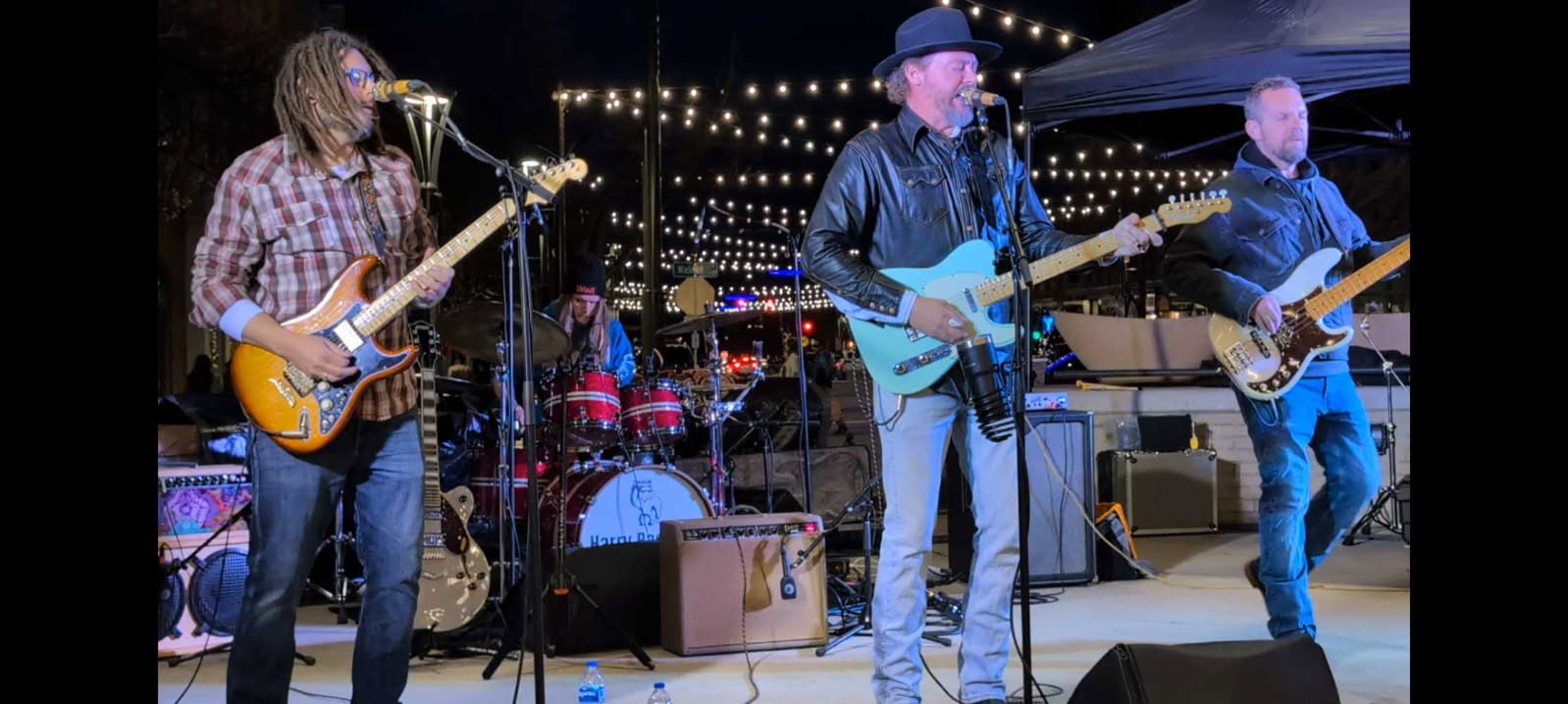 Four musicians perform on an outdoor stage with string lights above.