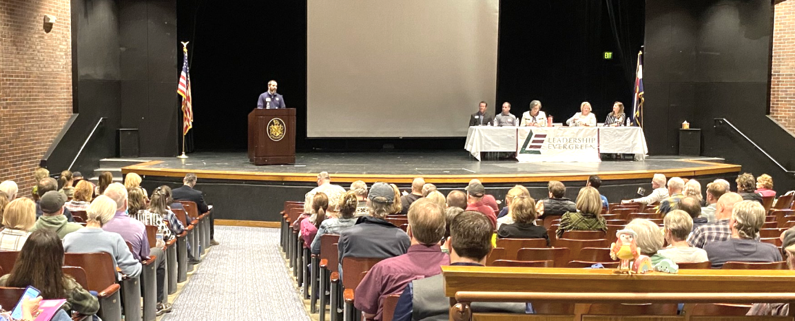 A speaker at a podium addresses an audience in a hall. A panel of five people sits at a table on the stage under a "Leadership Evergreen" banner.