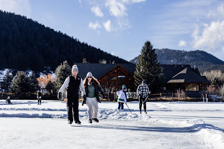 People ice skating outdoors by a cabin, mountains in the background.