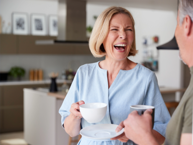 A woman laughs joyfully while holding a cup and plate, engaging with a man in a bright kitchen setting.