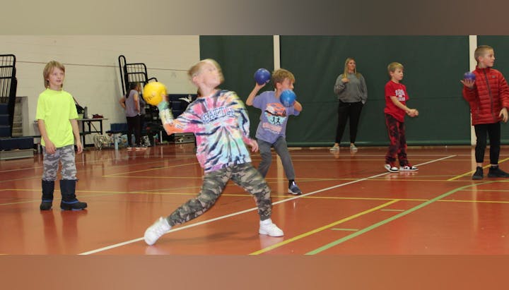 Children playing in a gymnasium, some throwing balls; an adult supervises in the background.