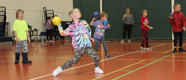 Children playing in a gymnasium, some throwing balls; an adult supervises in the background.