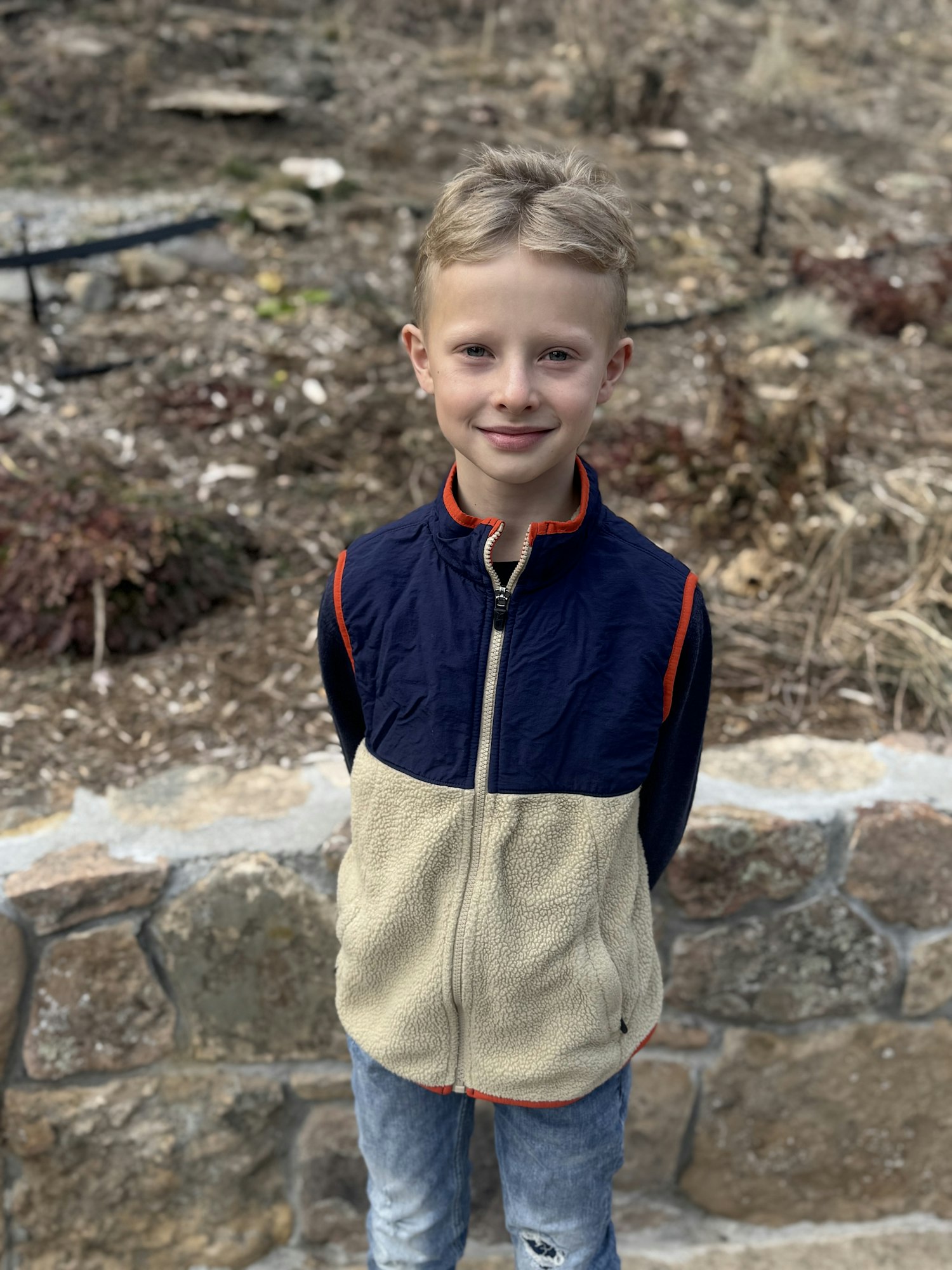 A child standing outside on a stone path, wearing a blue and beige jacket with hands behind the back, smiling.