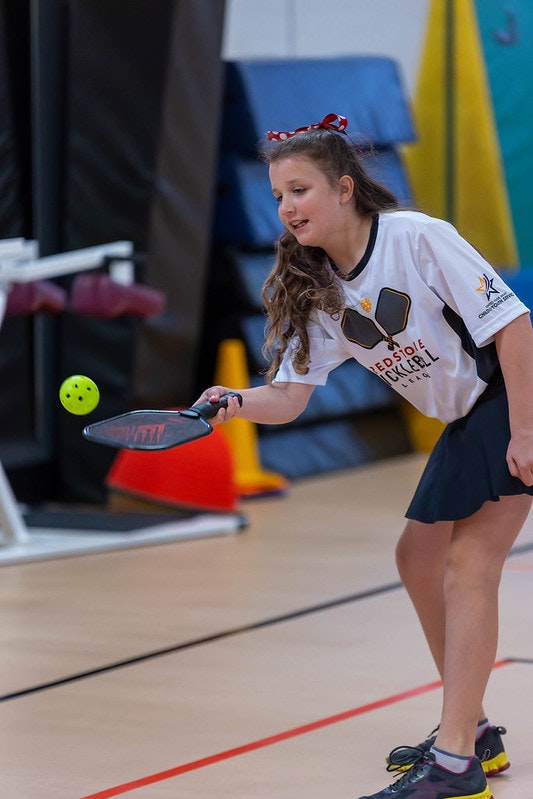 A girl playing pickleball indoors, wearing a white shirt, and hitting a ball with a paddle.