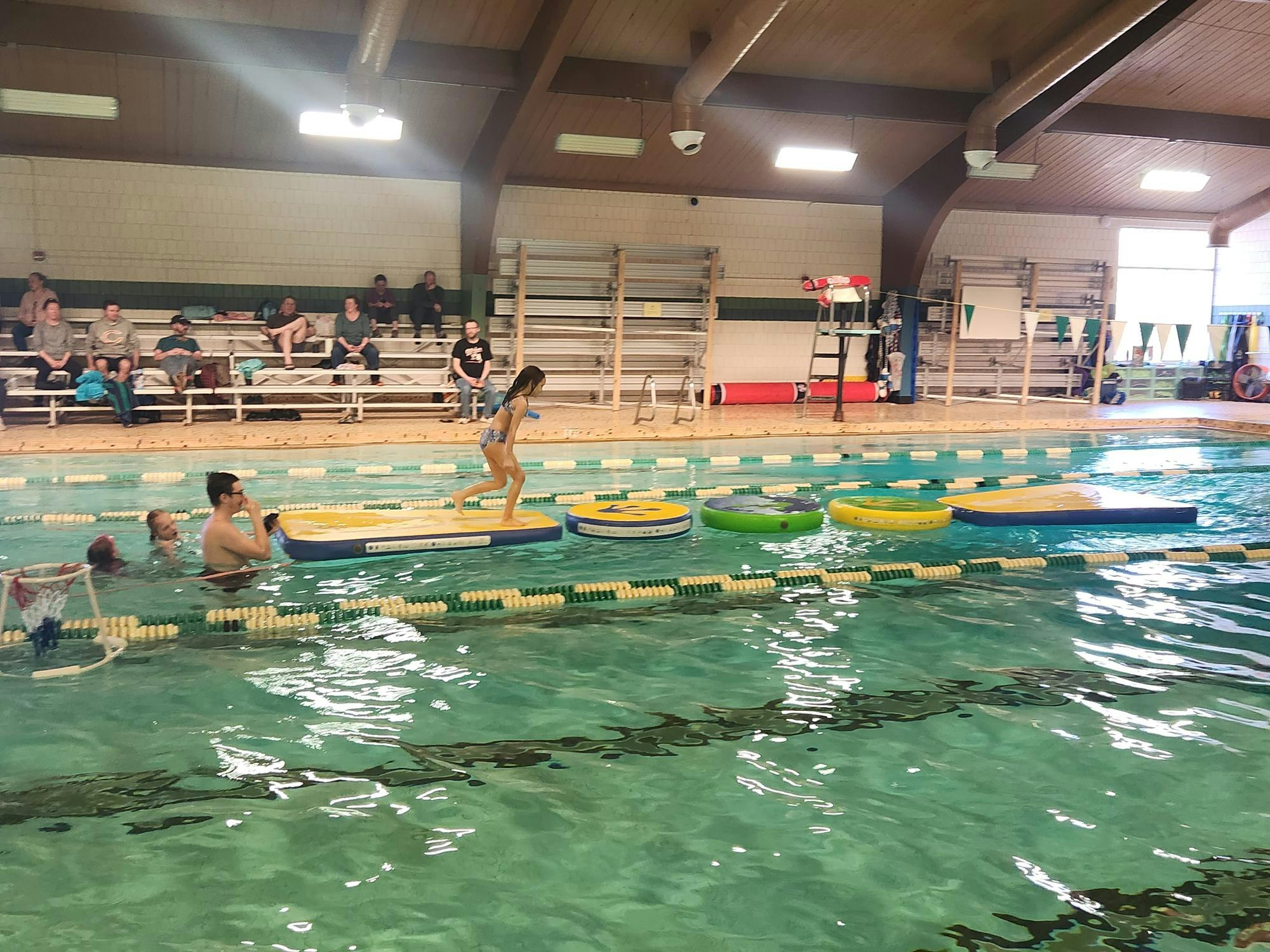 An indoor swimming pool with people, floating rings, and spectators on benches.