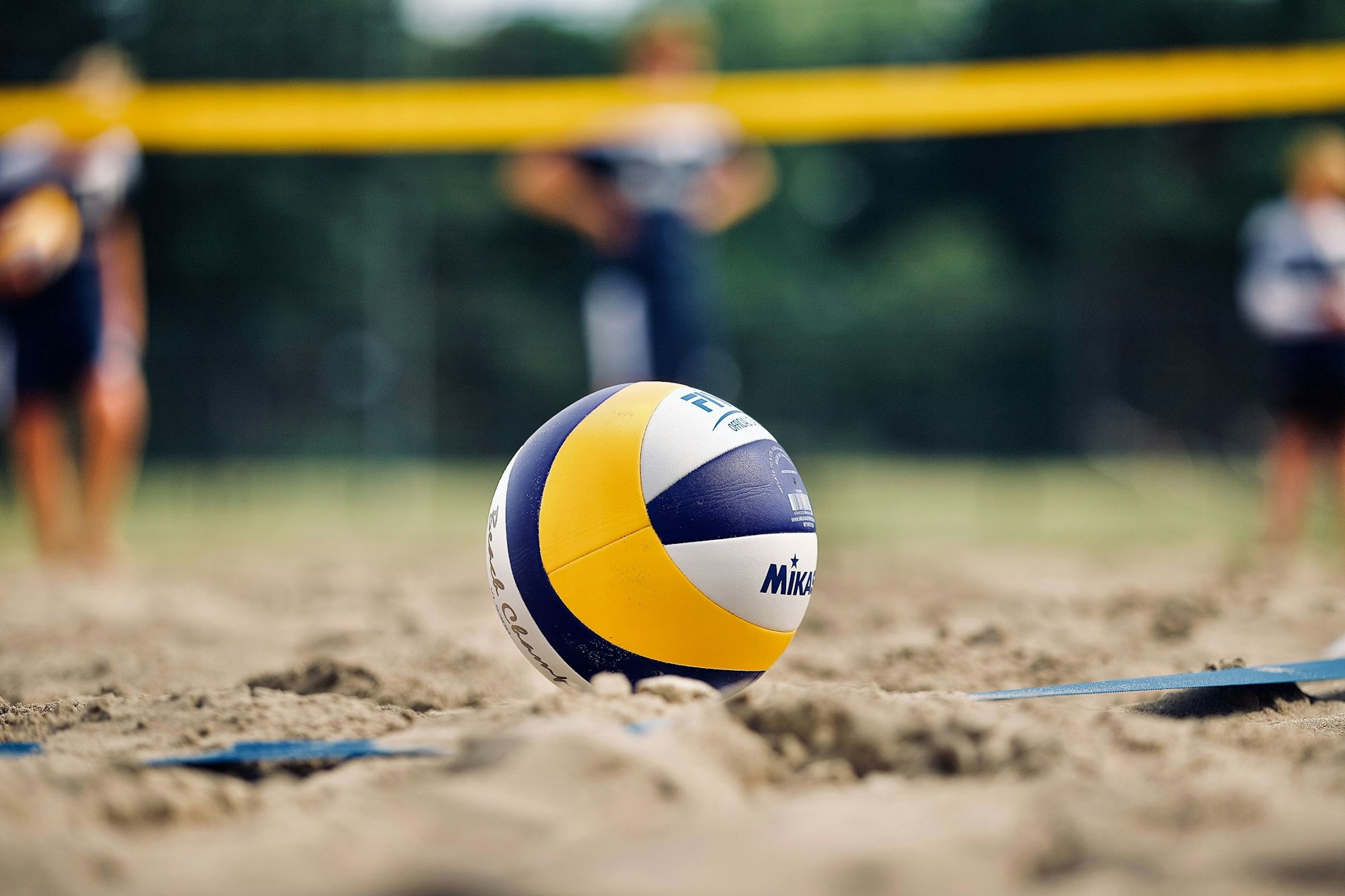 A beach volleyball on sand with blurred players and a net in the background.