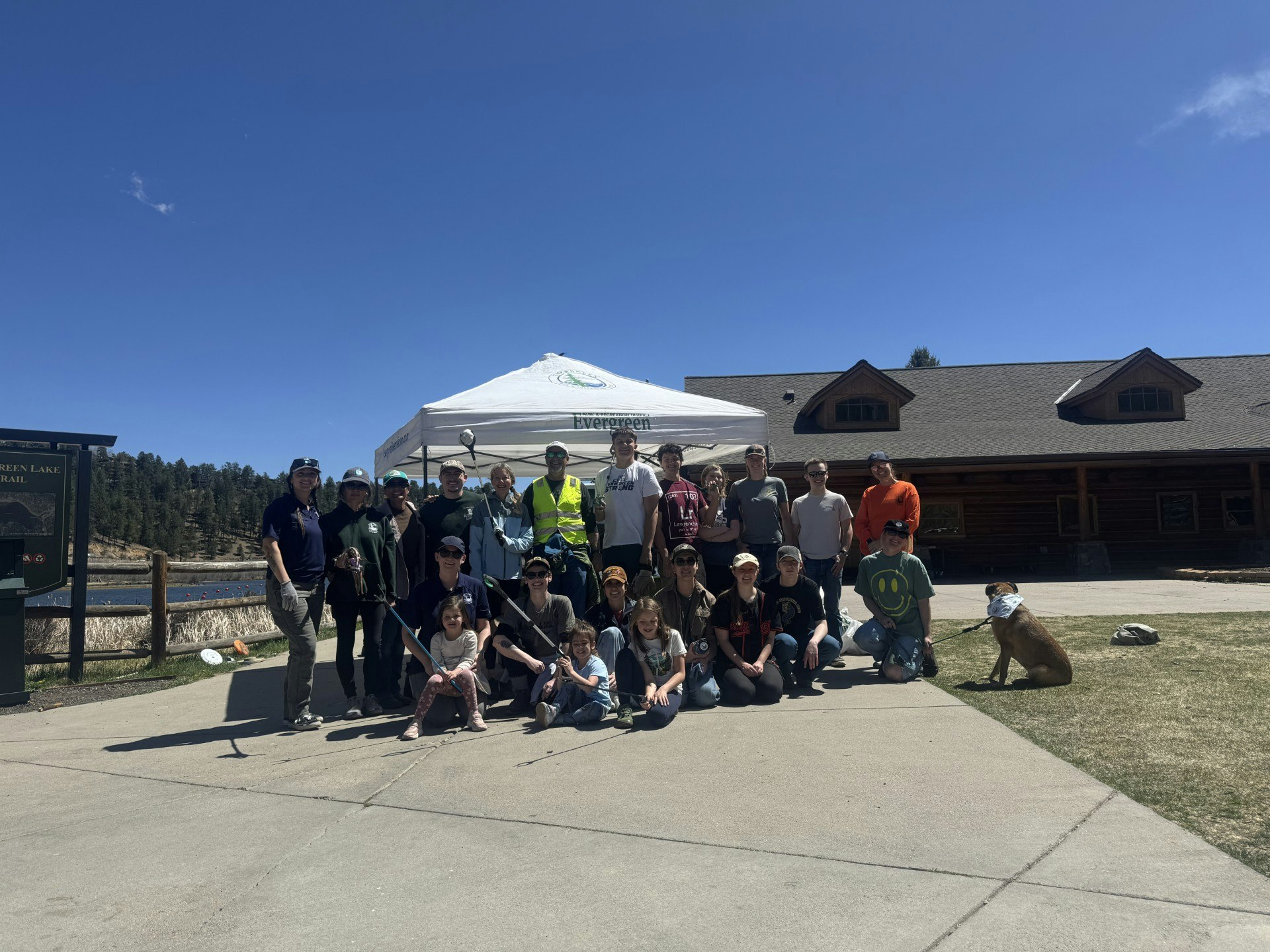 A group of people, including adults and children, pose together outdoors near a lodge, with a dog and a tent in the background.