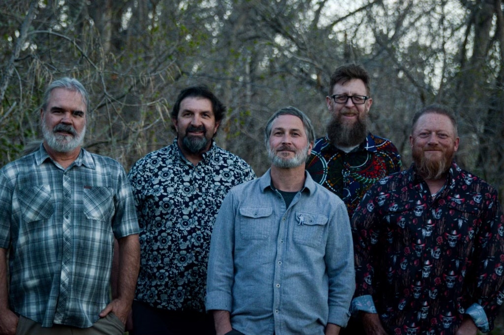 A group of five men standing together outdoors, wearing various patterned shirts, with trees in the background.