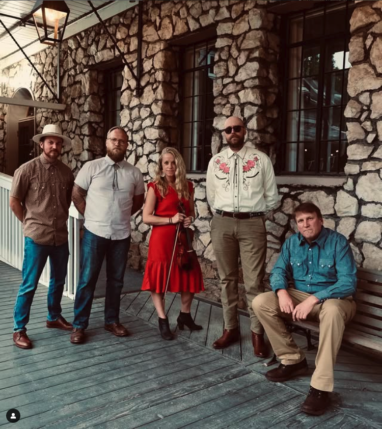 Five people stand outside a stone building, one holds a violin.