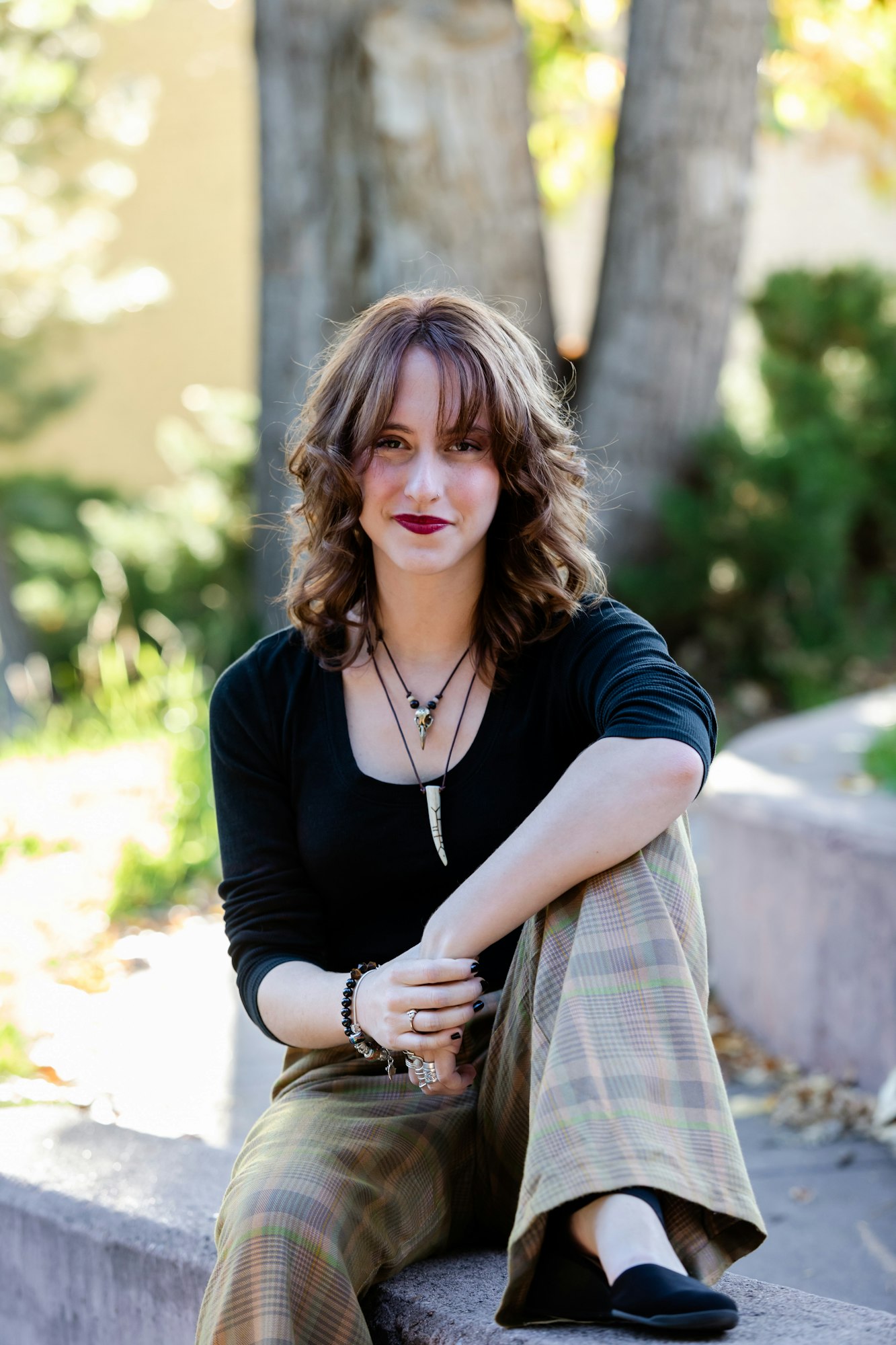 A person with curly hair sitting outdoors on a ledge, wearing plaid pants and a black top, accessorized with necklaces and bracelets.