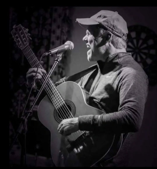 A person is passionately playing a guitar and singing into a microphone, wearing a cap.