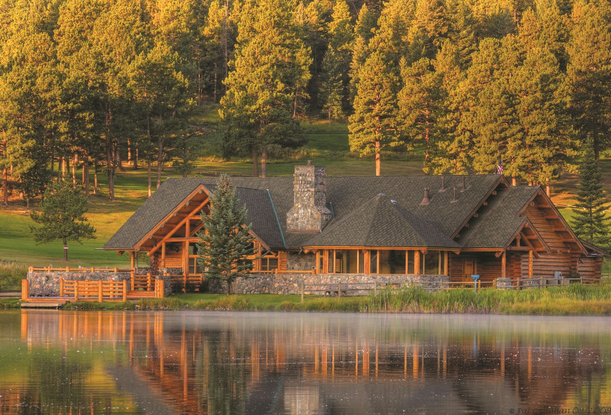 A serene log cabin by a lake with a backdrop of lush green pine trees bathed in warm sunlight.