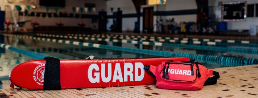 Lifeguard buoy and bag marked "GUARD" on a poolside with lanes in the background.