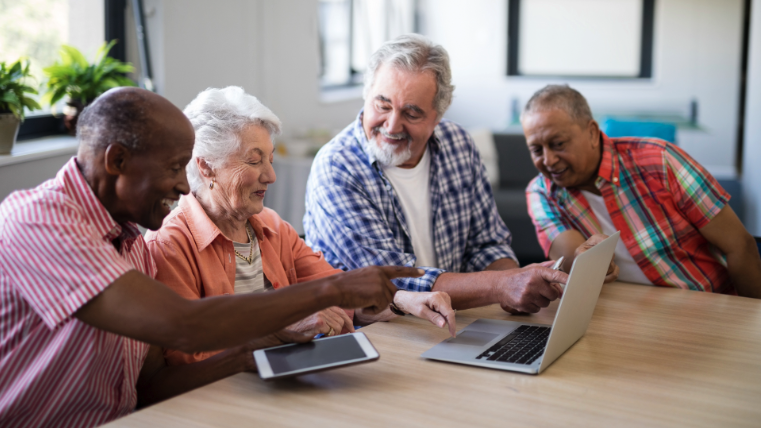 Four seniors happily engaged with a laptop and tablet at a table, sharing a moment.