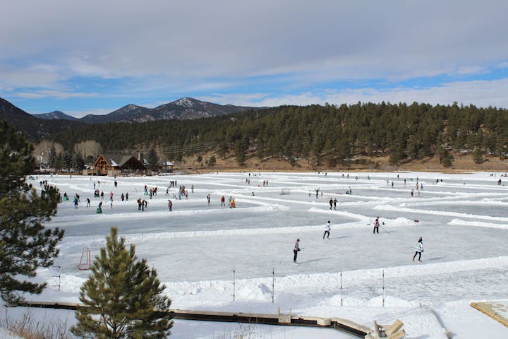 Outdoor ice rink with skaters, surrounded by snow, pine trees, and mountains in the background.