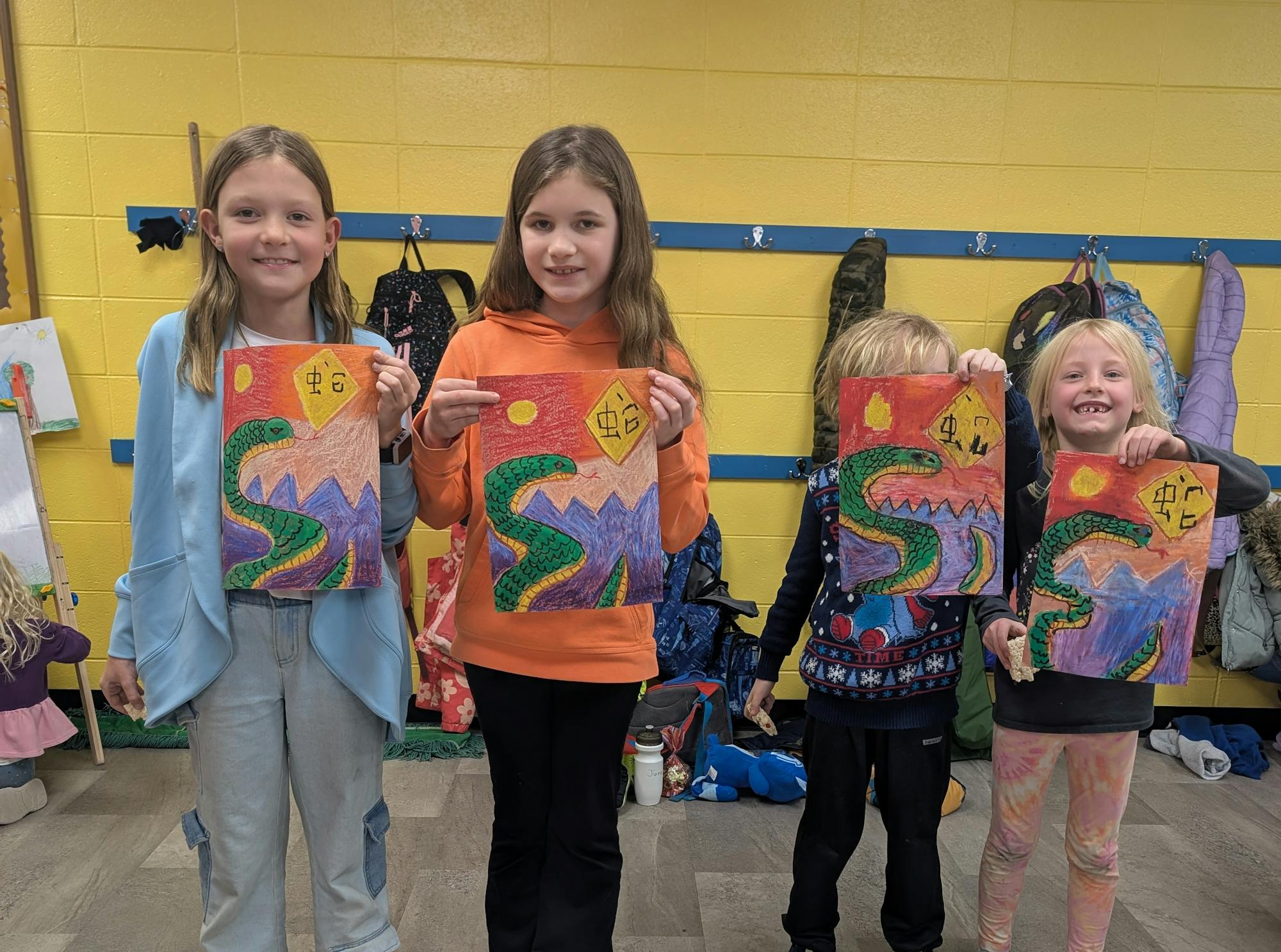 Four children hold colorful drawings featuring a dragon-like creature. The background is yellow, and they are in a classroom setting.