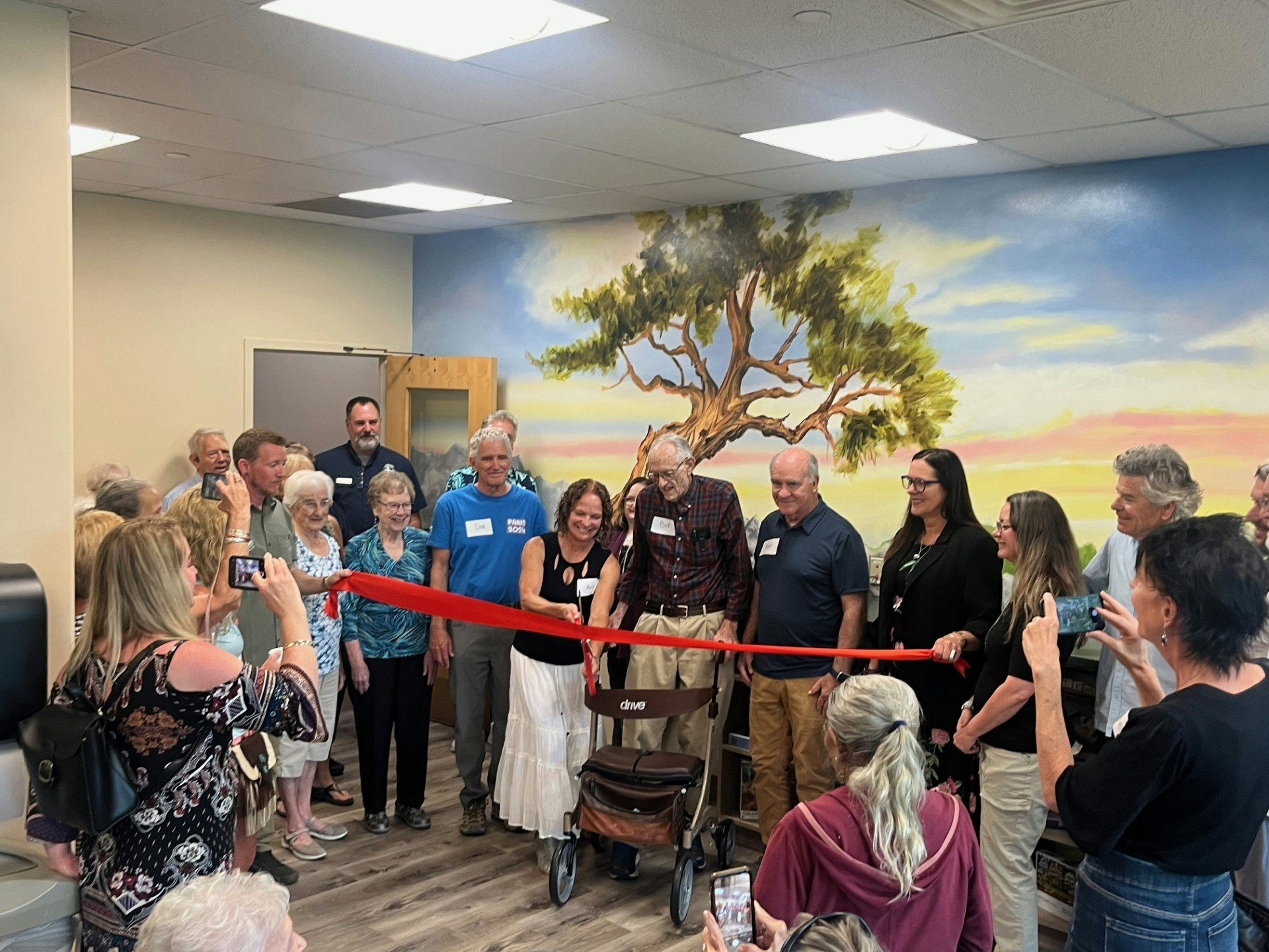 A group of people at a ribbon-cutting ceremony indoors, with a mural of a tree in the background.