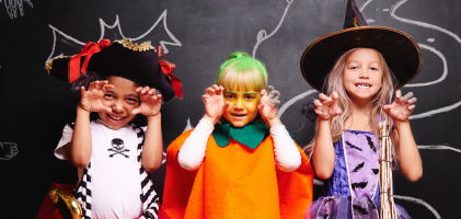 Three children in Halloween costumes: a pirate, a pumpkin, and a witch, posing playfully against a chalkboard backdrop.