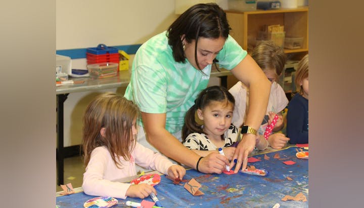 A teacher assisting children with crafts at a cluttered table.