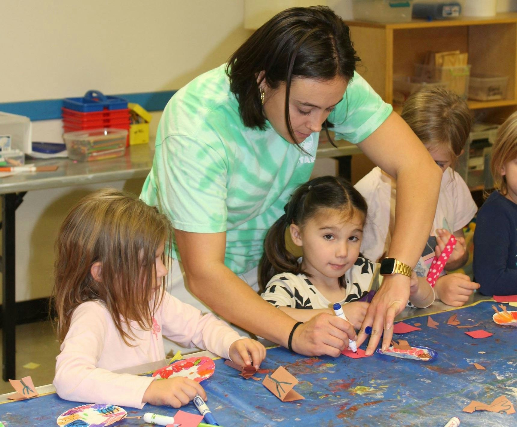 A teacher assisting children with crafts at a cluttered table.