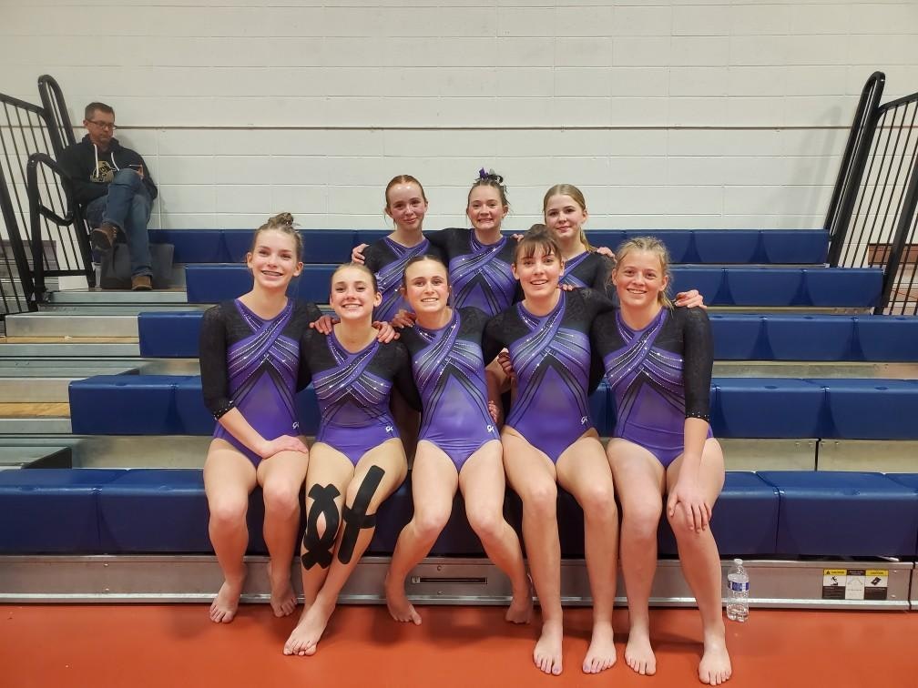 A group of gymnasts in purple uniforms sitting on bleachers, smiling for a photo.